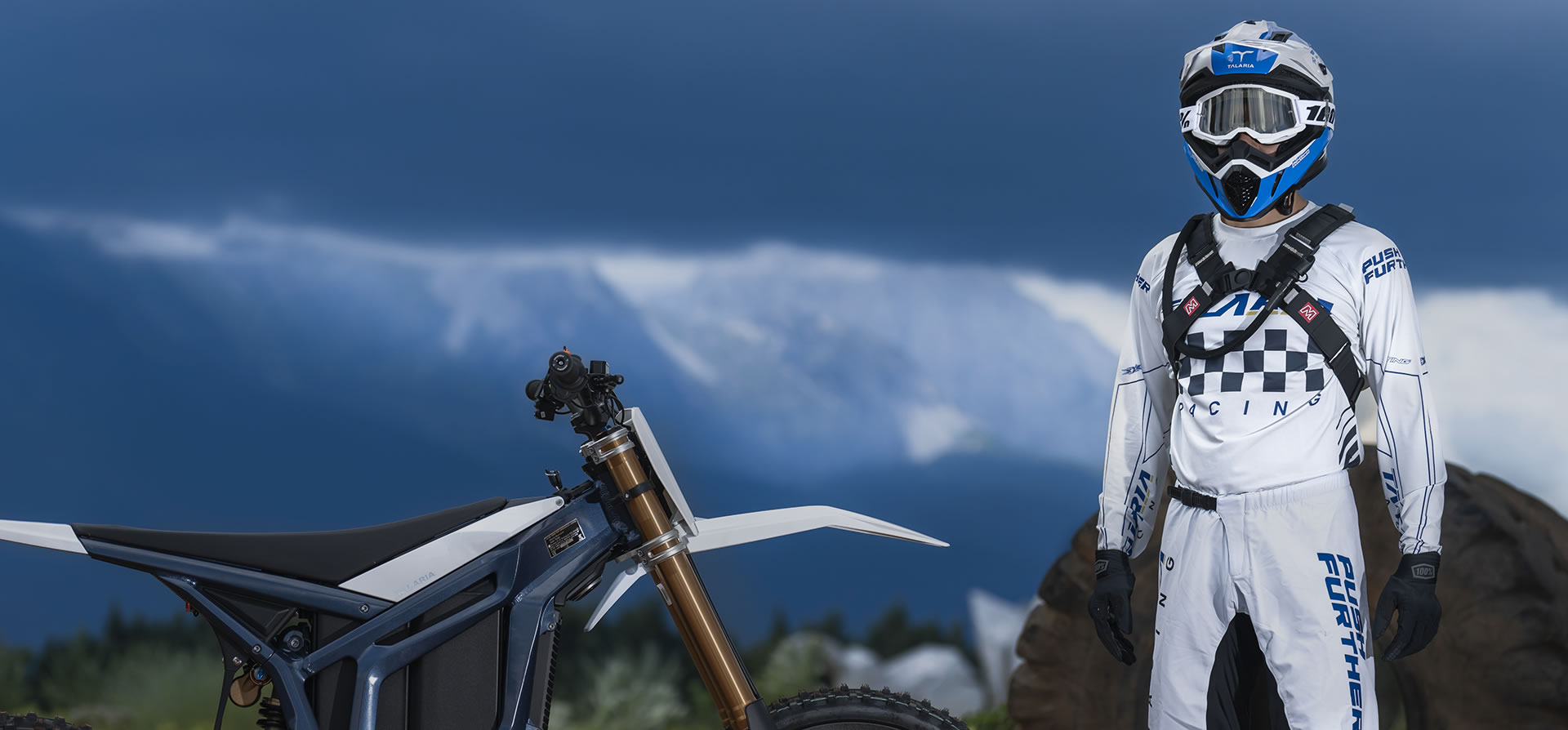 A rider in white gear standing beside a Talaria motorcycle under a stormy sky